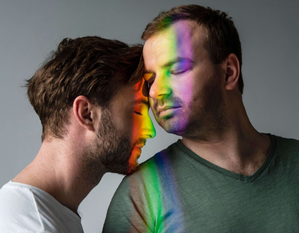 A tender moment between two men with their foreheads touching, eyes closed, and a rainbow light cast over their faces. This powerful photo captures love, connection, and the importance of mental health and emotional support within the Trans and LGBT+ communities, celebrating pride, positivity, and equality.