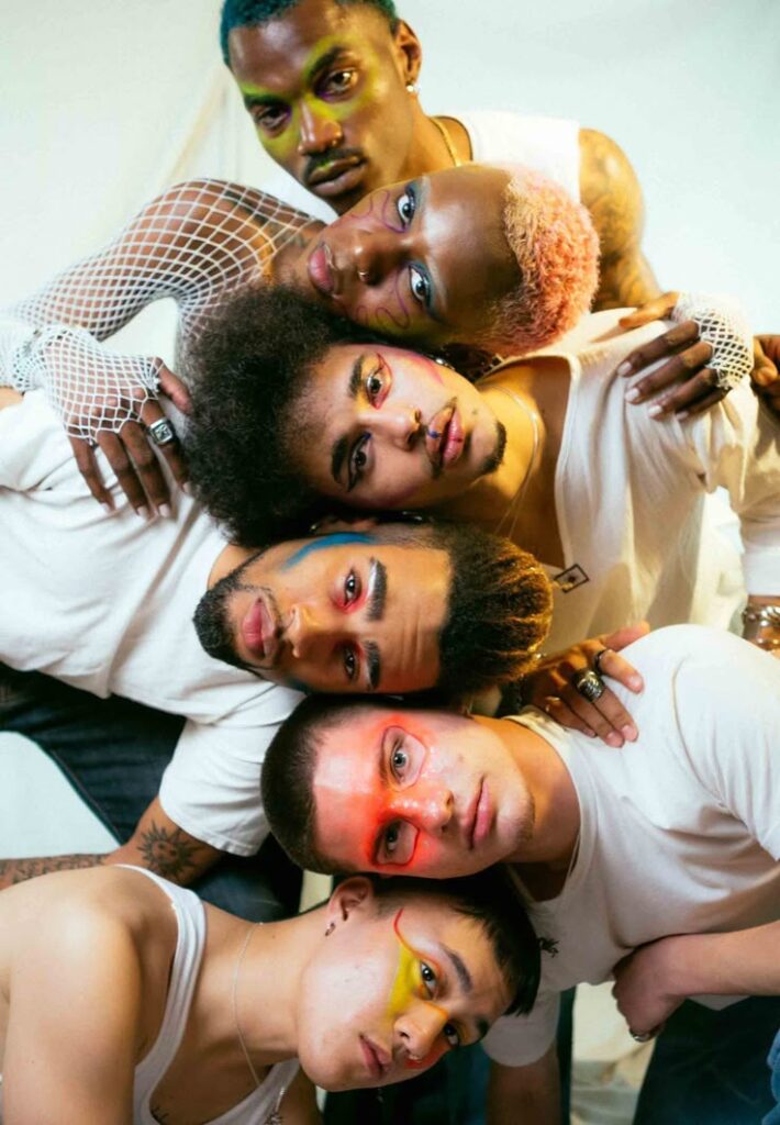 Six gender-diverse individuals with bold makeup and artistic styling, closely grouped and embodying fluid and non-conforming gender identities.