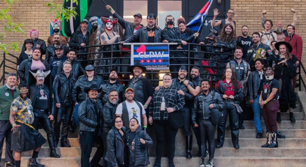 A large group of leather pride community members gathered together for a photo at an outdoor event, many wearing leather jackets, vests, and caps, representing the diversity and solidarity of leather culture.
